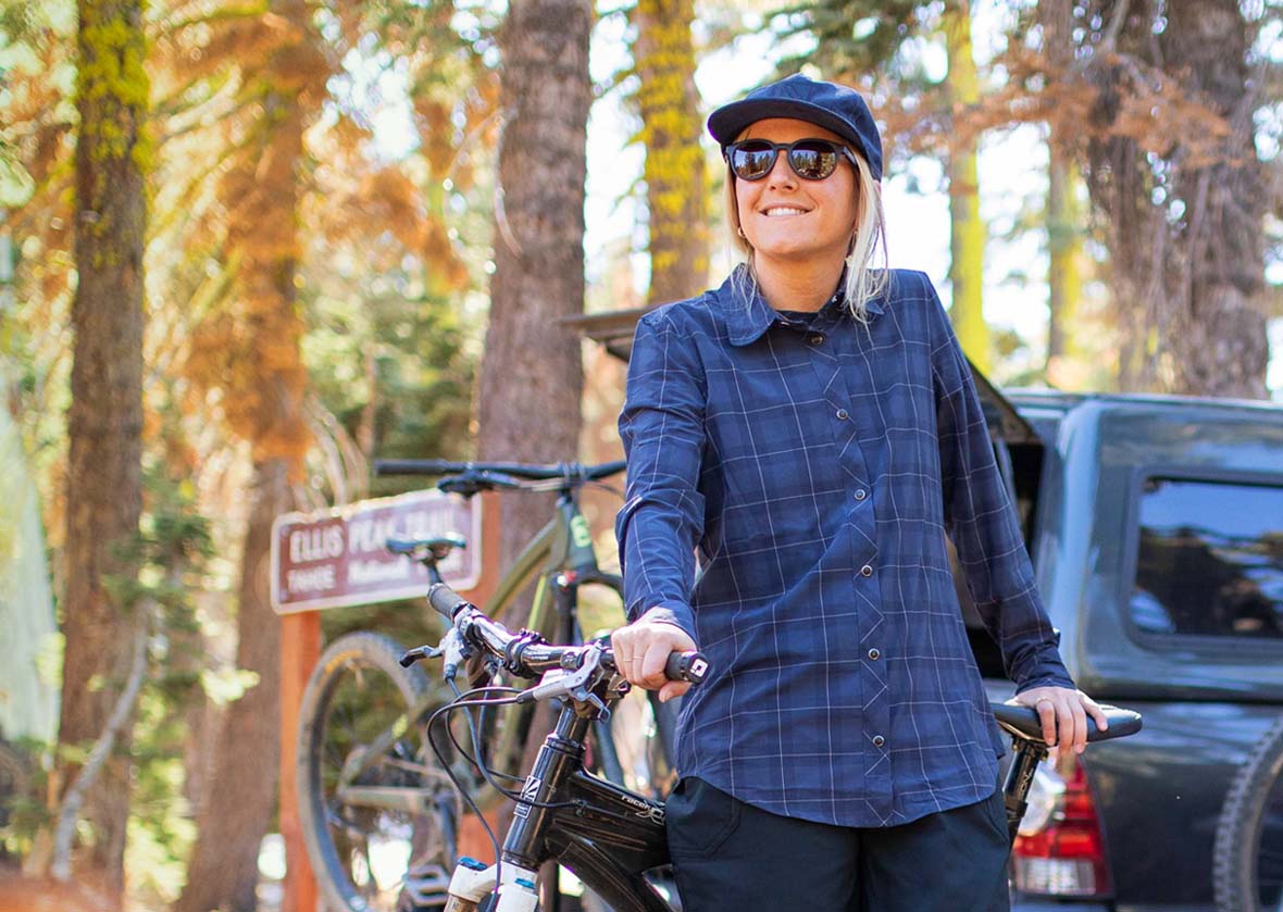 Woman wearing a blue plaid shirt with sunglasses and a hat, standing next to a bike