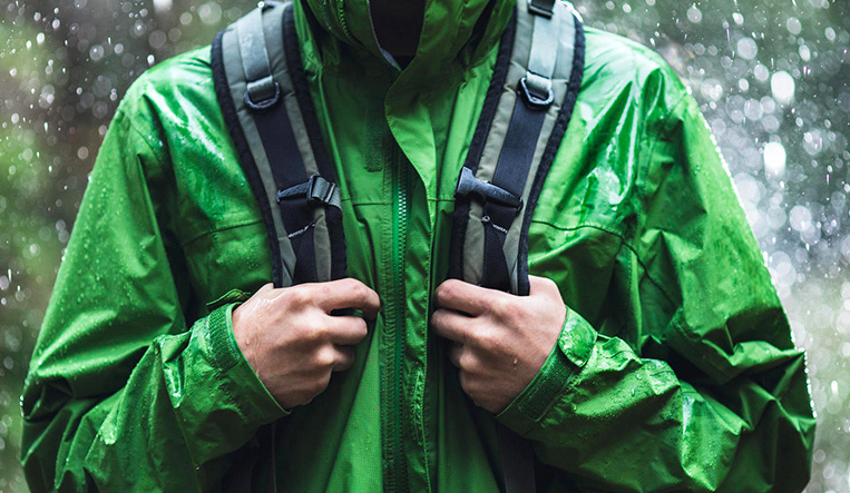 Person in green raincoat holding backpack straps in the rain, face not in view